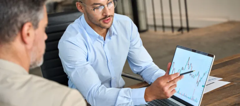 Two men sit in front of a tablet screen, looking at financial trends that are projecting in an upwards direction