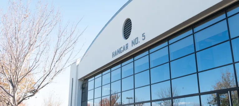 A large, white building with blue glass windows and a curved roof sits next to a leafless tree and in front of a blue sky, there is signage on the building that reads "Hangar No.5"