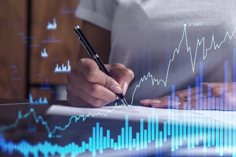 Image of a woman's hand writing notes with a stock ticker reflected on a screen in front to her