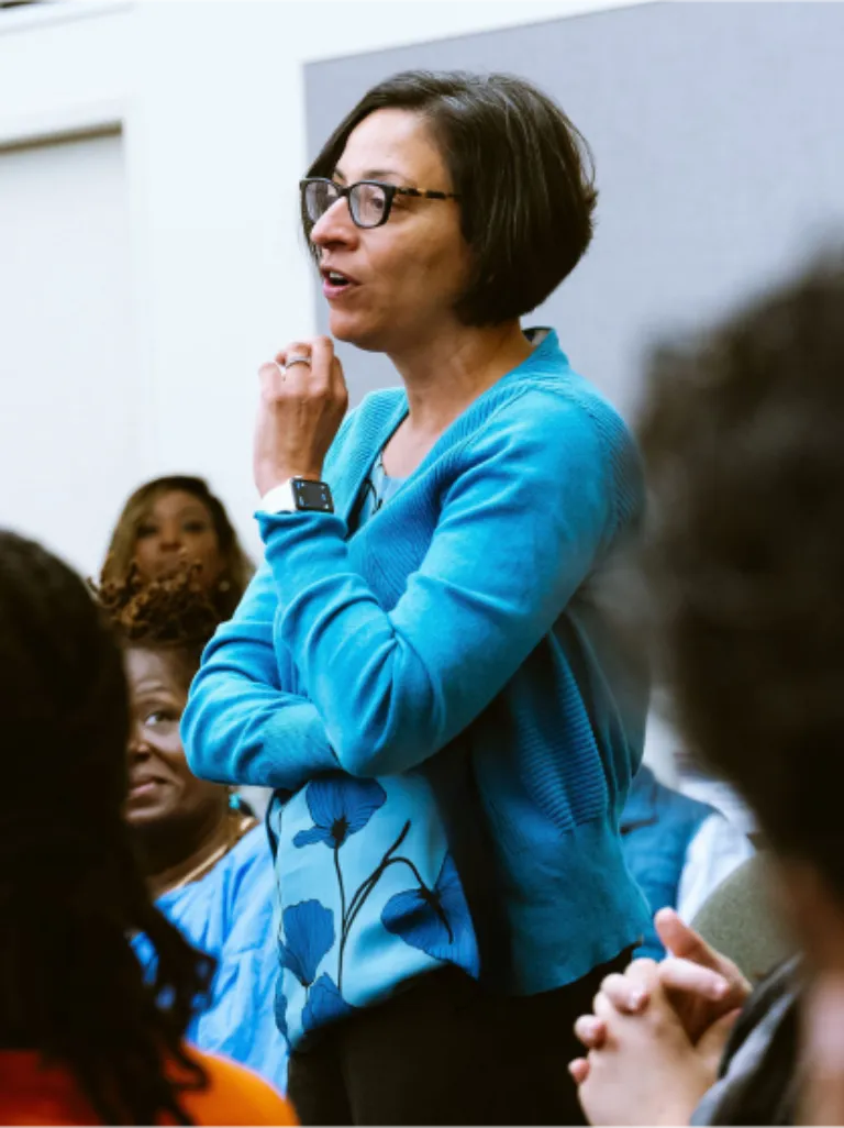 A woman in a blue sweater and blouse stands amongst a crowd of people talking as they look up to her