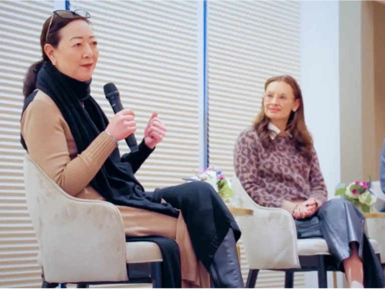 Rhea Suh sits in a chair with a microphone in her hand while a woman in the foreground looks turned to her