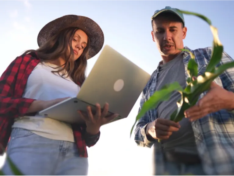 A woman in a sunhat holds a laptop and takes notes while speaking to a farmer holding one of his crops