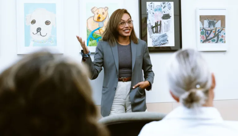 A woman gives an engaging presentation in front of an audience, gesturing confidently. Behind her are framed animal artworks, creating a casual and creative atmosphere.