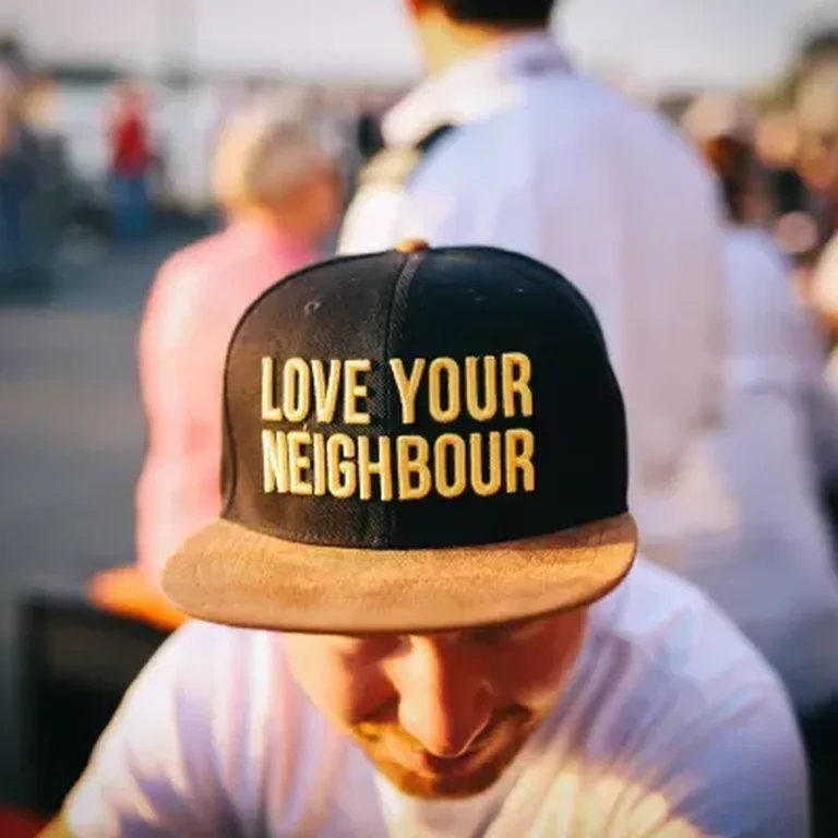 Person at an outdoor event looking down wearing a baseball cap with "Love Your Neighbor" in yellow letters on black background