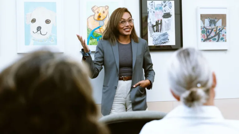 A woman gives an engaging presentation in front of an audience, gesturing confidently. Behind her are framed animal artworks, creating a casual and creative atmosphere.