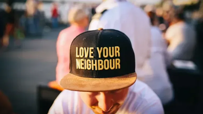 Person at an outdoor event looking down wearing a baseball cap with "Love Your Neighbor" in yellow letters on black background
