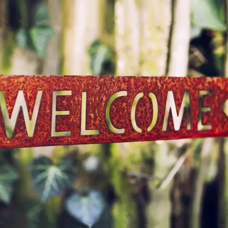 A rustic metal sign with the word "WELCOME" cut out is set against a background of lush greenery