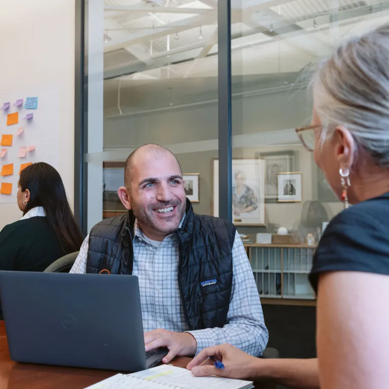 A man in a puffy black vest and checkered button-up shirt smiles at a woman with gray hair, earrings and a black short-sleeved shirt. They are sitting in an office setting.