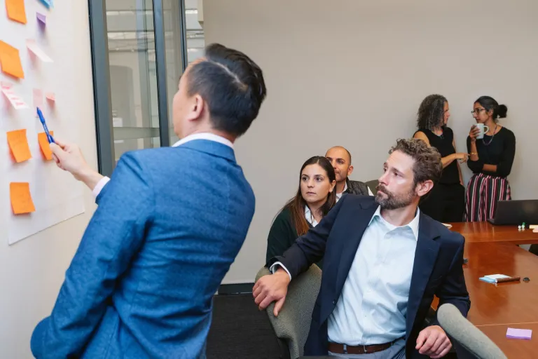 Group of people in a conference room looking at a facilitator who is placing post it notes on the wall