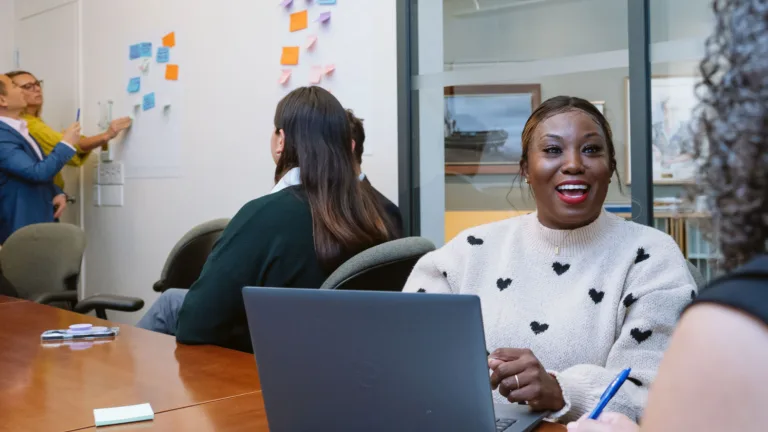 Two people at a white board at one end of the room with a woman looking on; one woman with a laptop laughing as another listens and takes notes