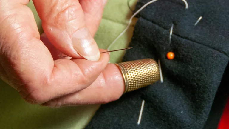 Close-up of a hand sewing fabric with a needle and thimble, pins in background