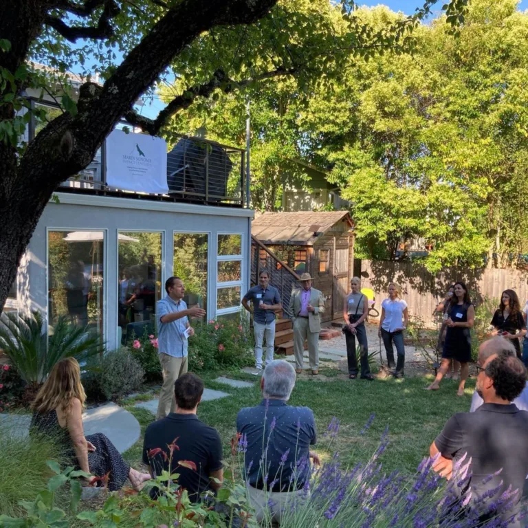 People gathered for an event in a backyard with trees and a modern building in the background.