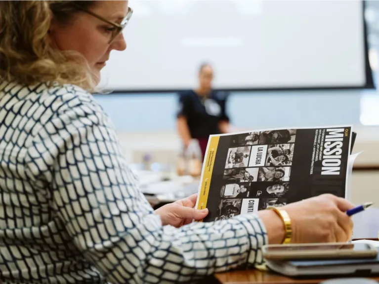 A woman with glasses and a white shirt flips through a magazine with a header that reads "Mission"