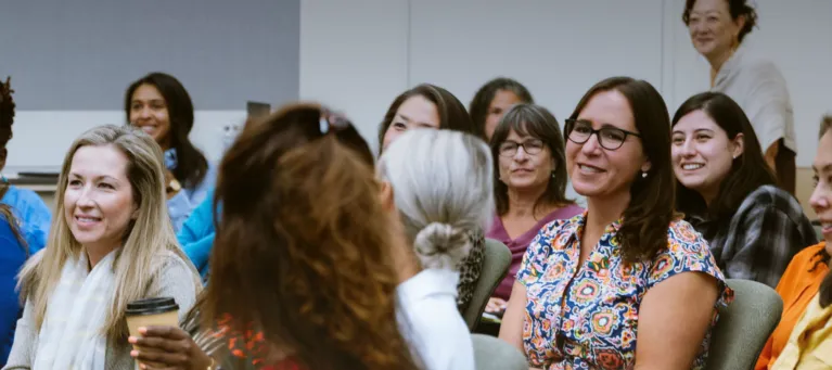 A diverse group of people talking amongst one another and smiling, with the focus being on a woman with glasses sitting in a chair