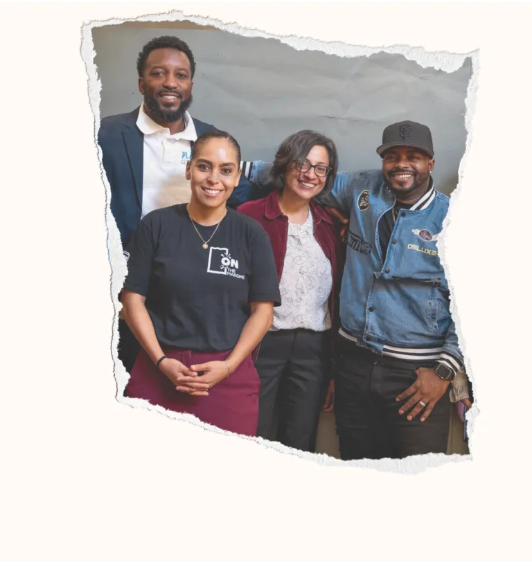 Four people, a well-dressed man in a suit, a woman in a black t-shirt and red pants, a woman in a red jacket and blouse, and a man in a black baseball cap and denim jacket stand smiling at the camera