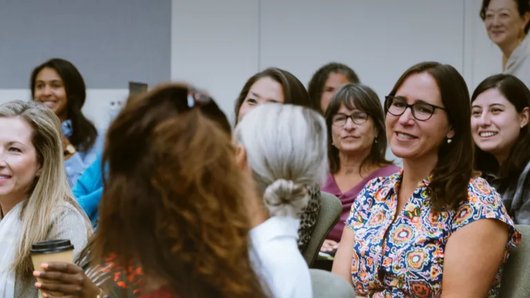 A diverse group of people talking amongst one another and smiling, with the focus being on a woman with glasses sitting in a chair