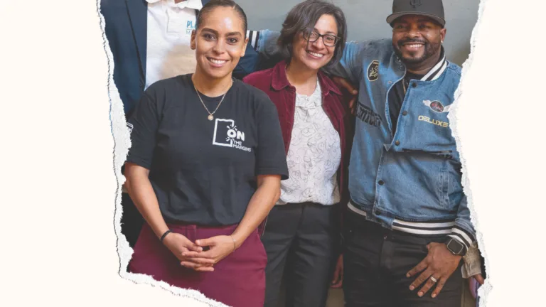 Four people, a well-dressed man in a suit, a woman in a black t-shirt and red pants, a woman in a red jacket and blouse, and a man in a black baseball cap and denim jacket stand smiling at the camera