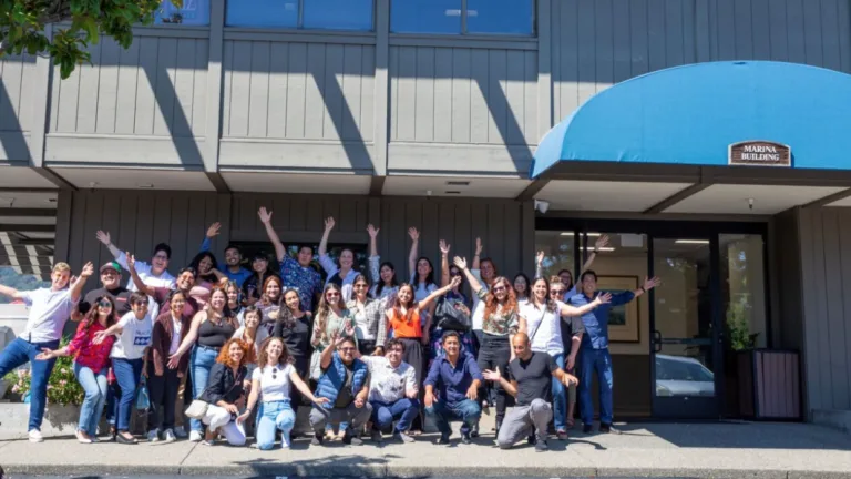 A group of people pose happily in front of a tan building with a blue awning