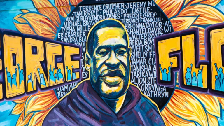 A mural of George Floyd at the center of a yellow and orange sunflower, with the names of other black persons and persons of color who have been victims of police brutality in the background.