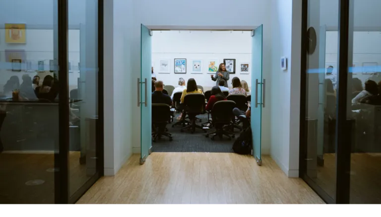 View through an open door of a meeting room with people seated, facing a presenter.