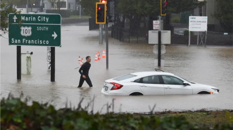 Man wading through a flooded street with a submerged car near San Francisco sign.