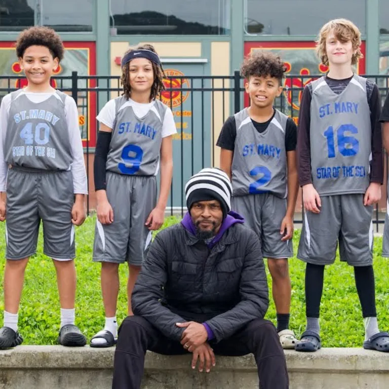 Group of children in basketball uniforms standing with a coach outdoors.