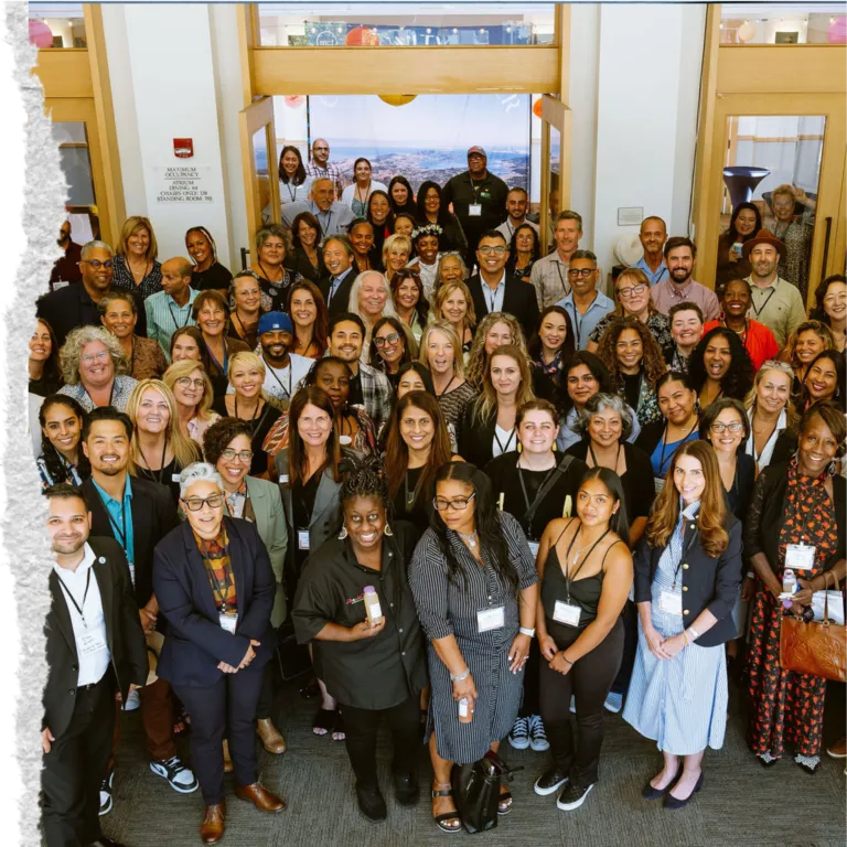 A large, diverse group of people stands in a big open room together, looking up and smiling for a group portrait