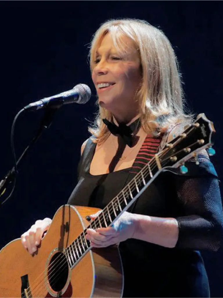 A woman musician in a black outfit playing an acoustic guitar on stage near a microphone.