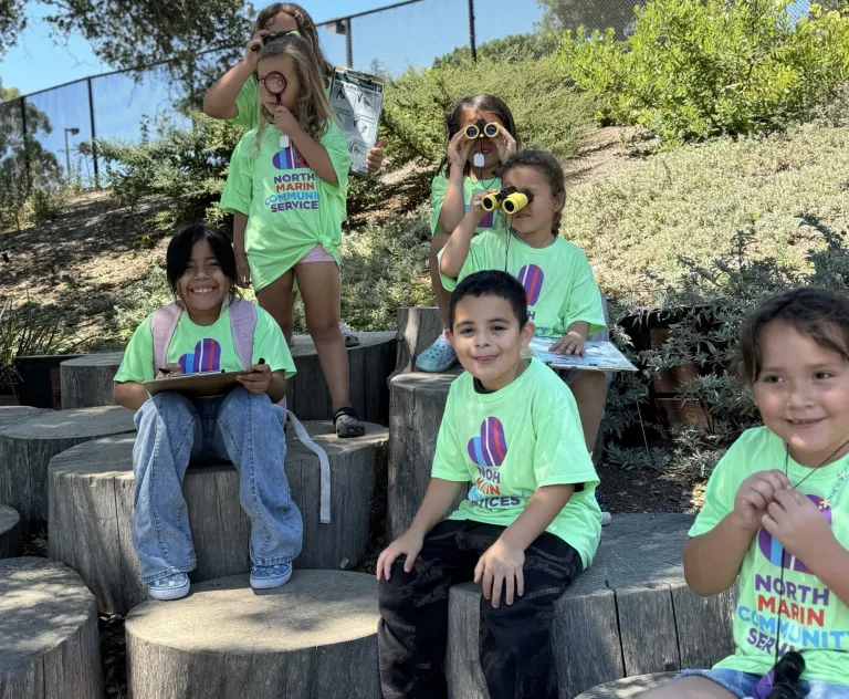 Image of a group of young children, sitting on logs, wearing t shirts from North Marin Community Services