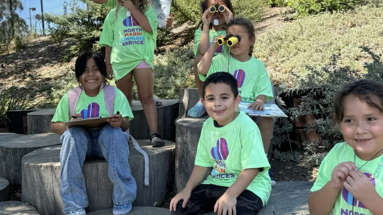 Image of a group of young children, sitting on logs, wearing t shirts from North Marin Community Services