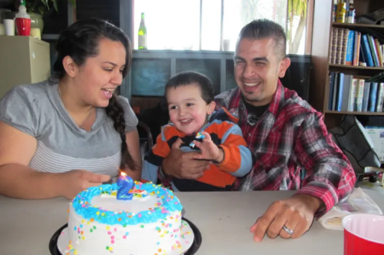 Mom, dad and child at a table with a birthday cake, with lit candles.