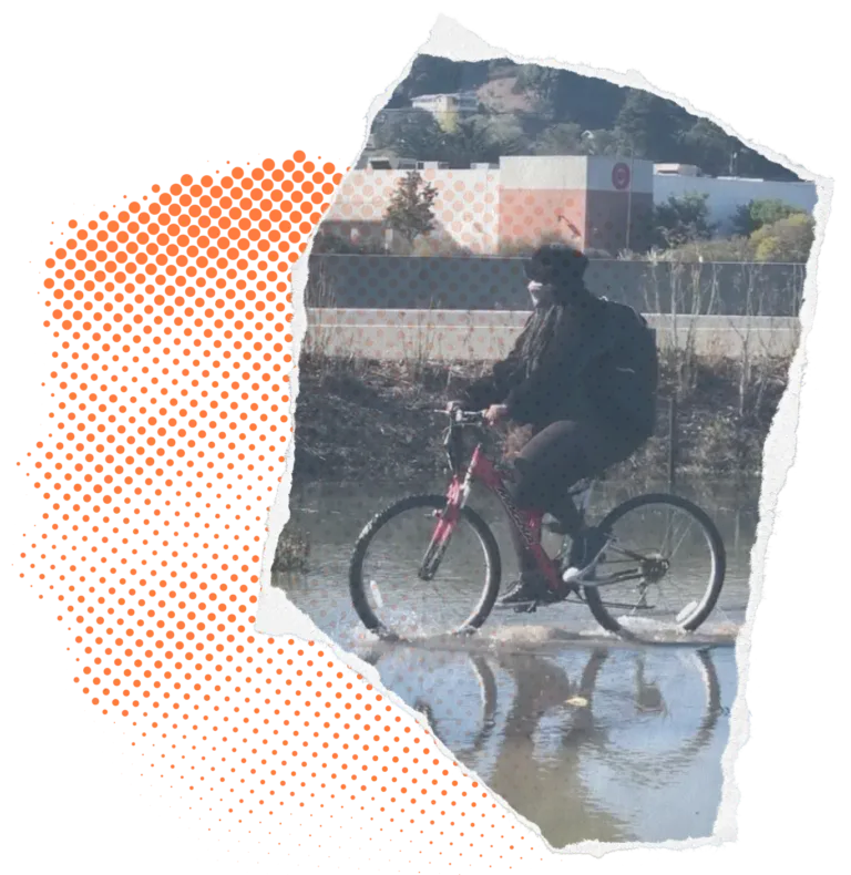 A person rides a bicycle through a shallow, flooded area, dressed in dark clothing with a backpack. The background features an industrial setting with distant hills and buildings. The image has a collage-style effect with torn paper edges and an orange halftone dot pattern on the left side,