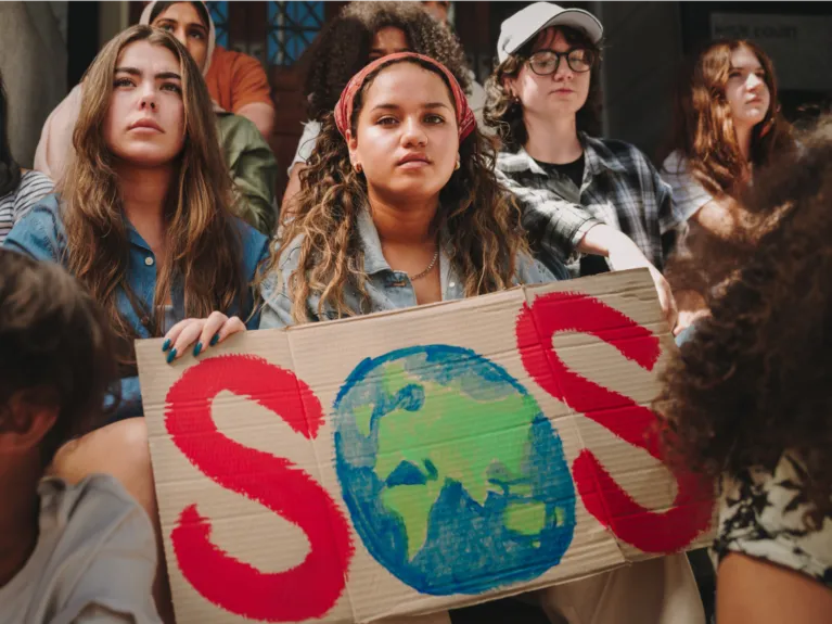 A group of teenagers and children of all demographics stand beside one another holding a cardboard sign that reads "SOS" in red paint with the O being a painting of the earth