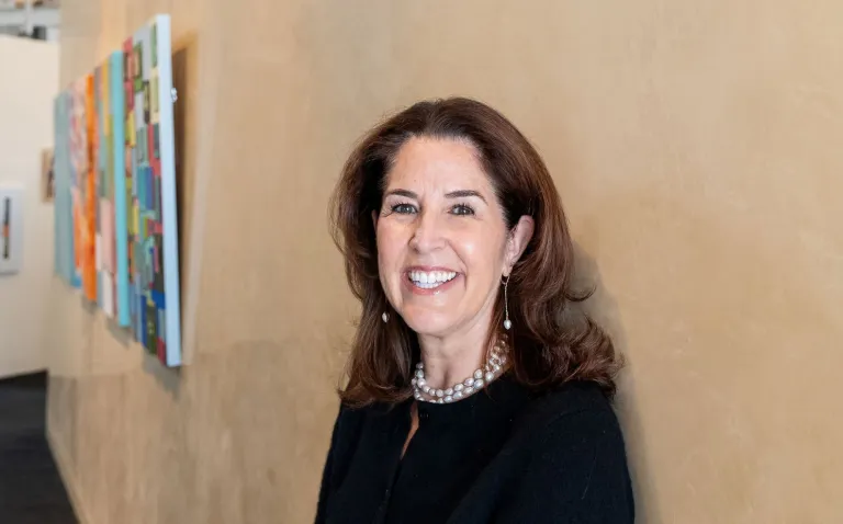 Photo of Janelle Cavanagh, smiling and leaning next to a cream wall; wearing a black sweater and pearls