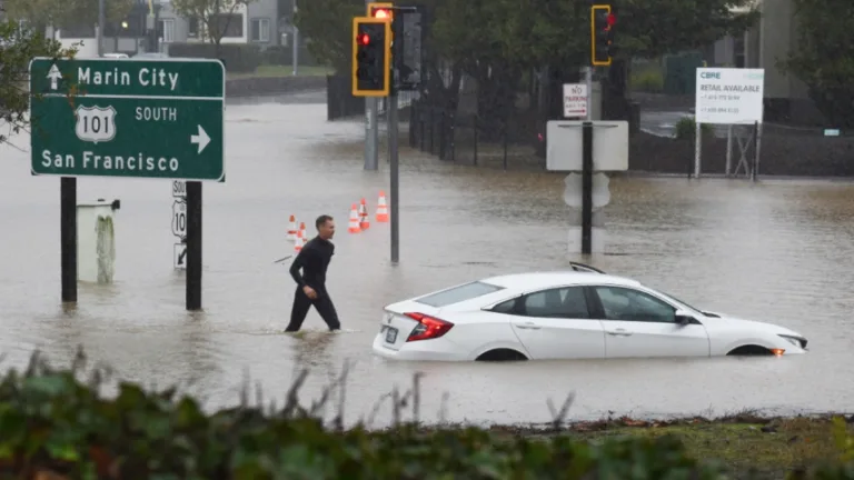 A man treads through knee-deep water next to a flooded car and a highway sign that reads "Marin City" and "101 South San Francisco"