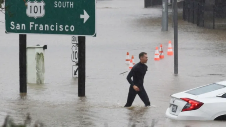 A man treads through knee-deep water next to a flooded car and a highway sign that reads "Marin City" and "101 South San Francisco"