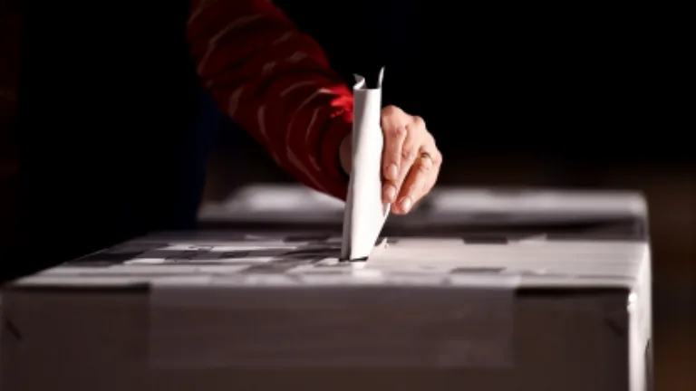 A person's hand is illuminated in light against a dark background while they stick their ballot into a box