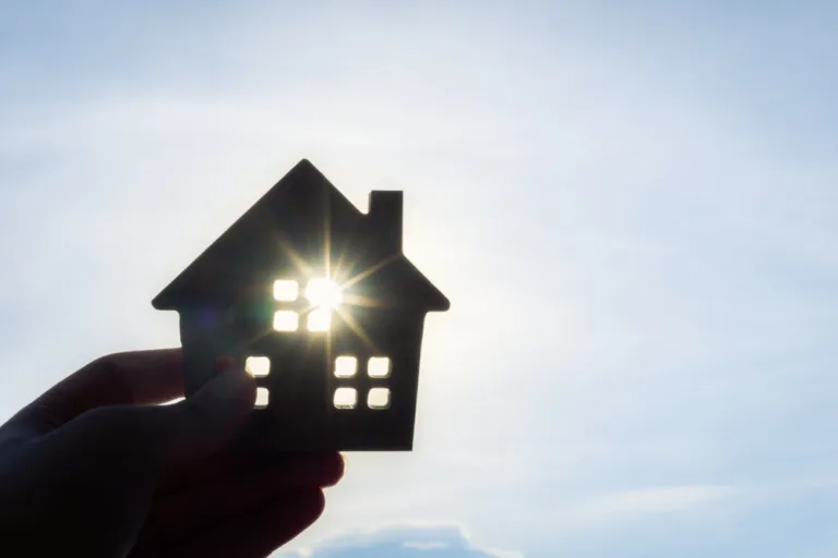 Silhouette of a hand holding a cut out of a house against the blue sky with the sun shining through one of the windows