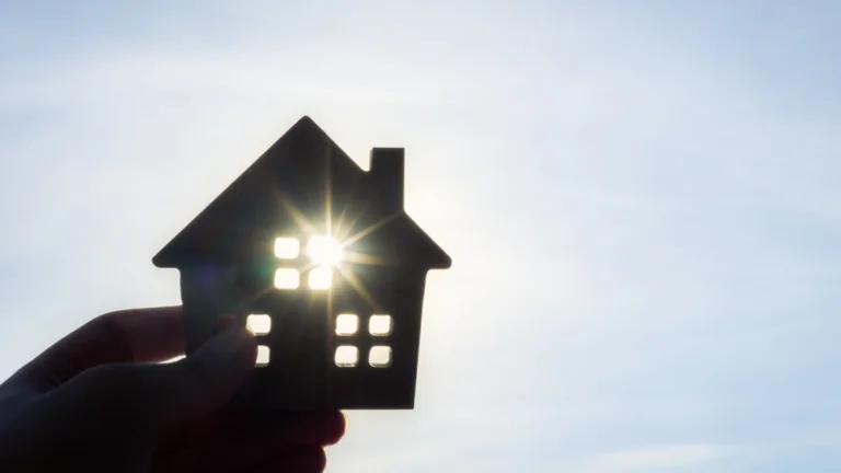 Silhouette of a hand holding a cut out of a house against the blue sky with the sun shining through one of the windows