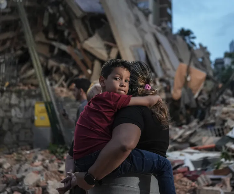 Image of a young boy being carried by a woman, only seeing her back, in a rubble