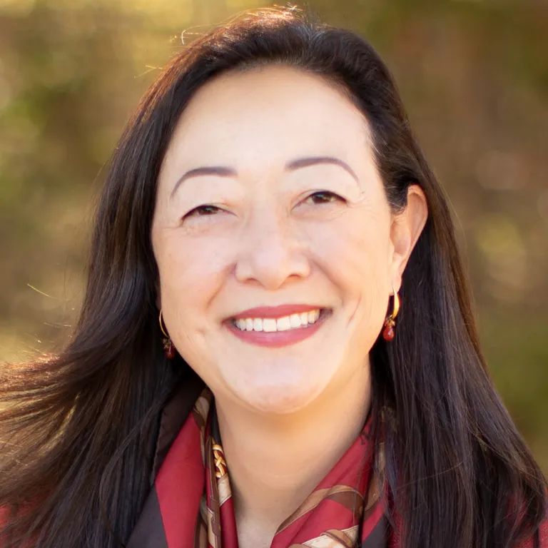 Headshot of Rhea Suh wearing a grey jacket, red blouse, and colorful scarf