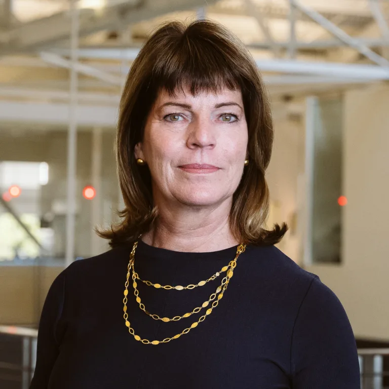 Ann P. Barber headshot in a navy dress with a gold chain necklace, standing indoors.