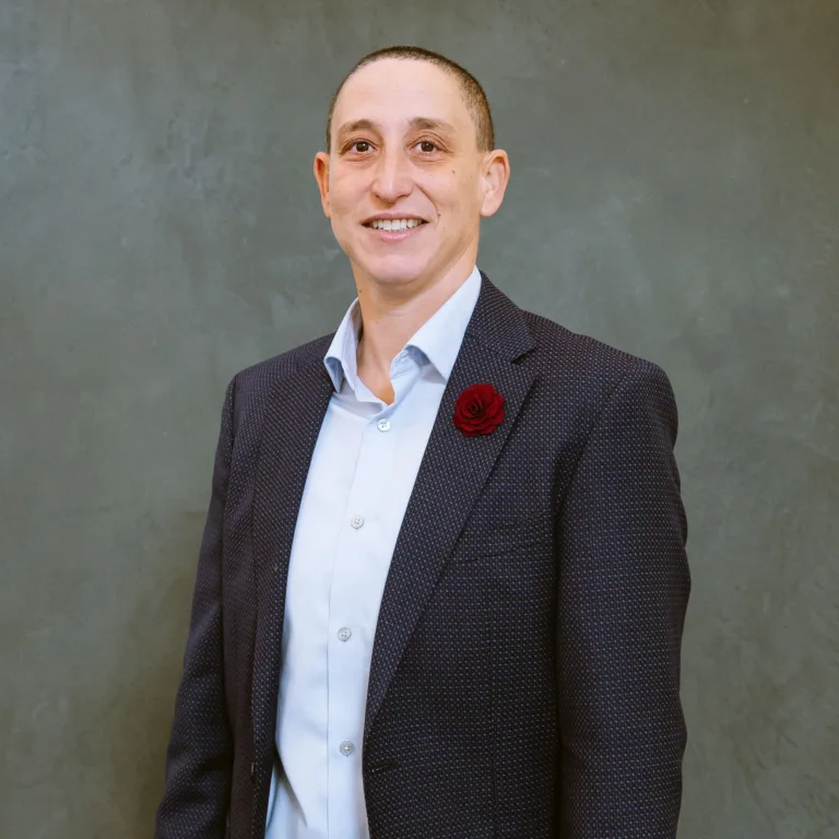 Headshot of Tara Medve in a patterned suit with a red boutonniere against a gray background.