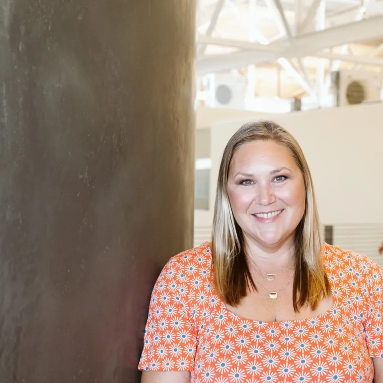 Image of Ashley Smith, leaning against a wall, smiling and wearing an orange and white top.