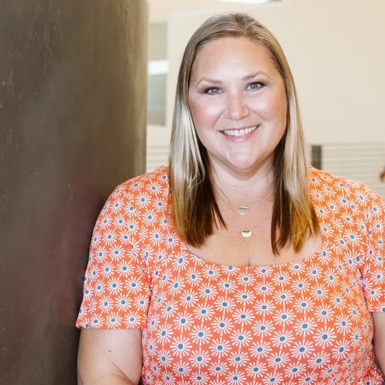 Image of Ashley Smith, leaning against a wall, smiling and wearing an orange and white top.