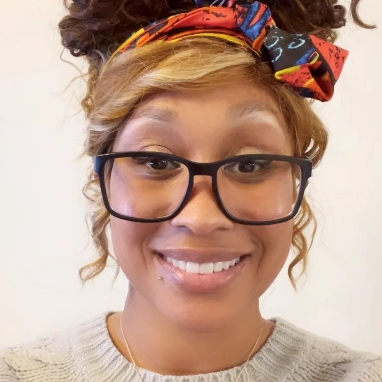 Head and shoulders image of Monique Bohman, smiling and wearing a colorful headscarf, black-rimmed glasses and a cream sweater