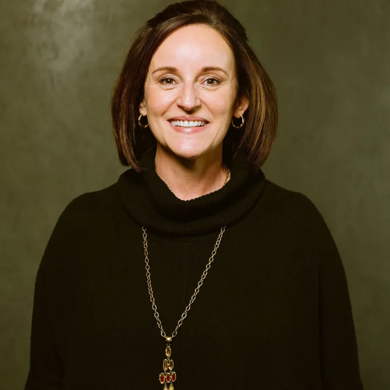 Image of Carmen Cruces smiling at camera, wearing a black turtle-neck sweater and a long gold chain.