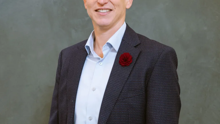 Headshot of Tara Medve in a patterned suit with a red boutonniere against a gray background.